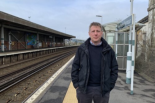 James MacCleary standing on the platform at Newhaven Harbour station. Behind him are empty tracks, station buildings and signalling paraphrenalia