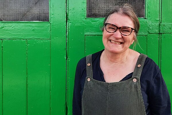 portrait shot of Edwina Livesey, dressed in dungarees, with glasses and brown hair tied back, against a set of green doors.