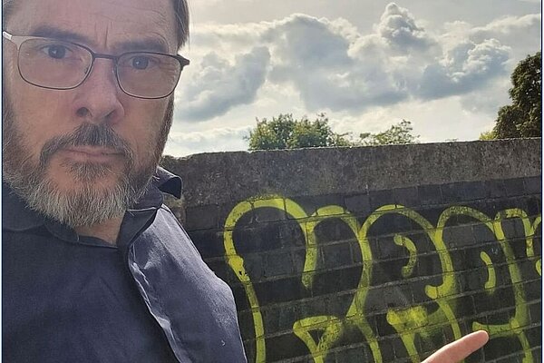 Kevin West in the foreground with glasses, dark hair and greying beard, points at grafitti on a bridge wall behind him.