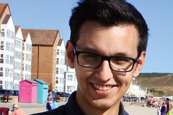 Head shot of Freddie, a young man with glasses and dark short hair. The background is a seafront scene including two colourful beach huts.