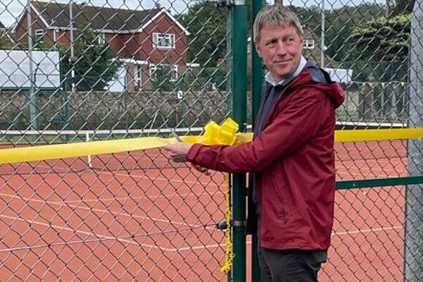 James opening new tennis courts in Seaford James is looking over his shoulder while holding scissors on a decoratively tied yellow ribbon across the gateway to a tennis court. He appears to be about to cut off his fingers.