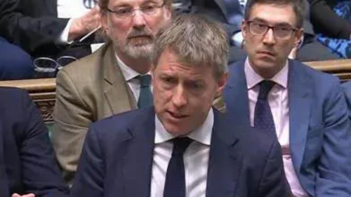 James, dressed in sober suit and tie, and with colleagues behind him, makes a point in the House of Commons