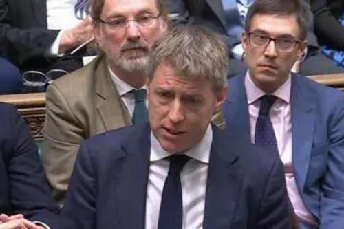 James, dressed in sober suit and tie, and with colleagues behind him, makes a point in the House of Commons
