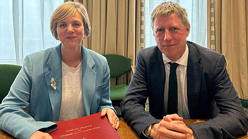 James seated at a table in front of some rather imposing curtains with Roads Minister Lilian Greenwood. She has an embossed folder in front of her.