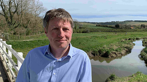 James MacCleary, in the foreground, on the South Downs Way near Alfriston, with a stream, fields, trees and blue sky behind him