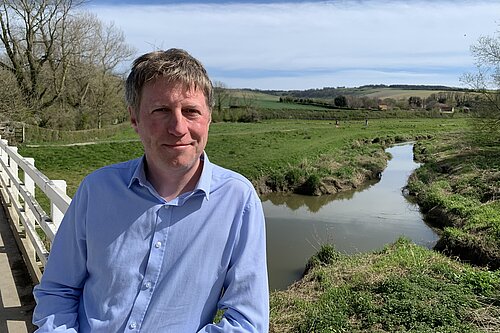 James MacCleary, in the foreground, on the South Downs Way near Alfriston, with a stream, fields, trees and blue sky behind him