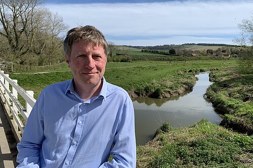 James MacCleary, in the foreground, on the South Downs Way near Alfriston, with a stream, fields, trees and blue sky behind him