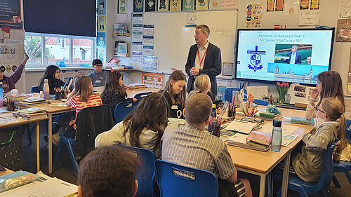 A busy classroom with a dozen children at tables and James at the front talking to them