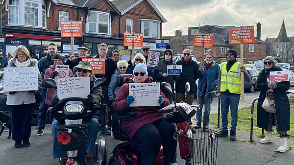 James MacCleary in a crowd of local residents holding placards protesting about uncontrolled parking in Polegate. At the front are two people in mobility scooters with handwritten signs saying "Please don't park across dropped kerbs". Several others are holding printed signs saying "Uncontrolled parking is destroying our town".