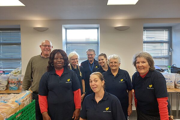 Councillors Janet Baah and John Lamb with six members of staff of Malling Foodbank. They are wearing Malling Foodbank t-shirts. On nearby tables are trays of bread and bags of food.