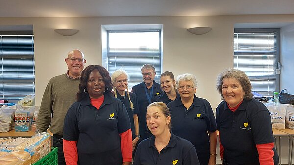 Councillors Janet Baah and John Lamb with six members of staff of Malling Foodbank. They are wearing Malling Foodbank t-shirts. On nearby tables are trays of bread and bags of food.