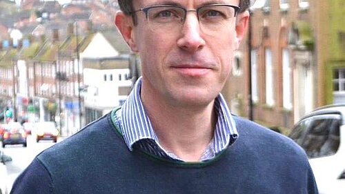 Close up of Ben with LibDem rosette, looking at the camera, with Lewes's high street in tha background