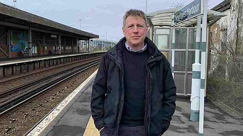 James MacCleary standing on the platform at Newhaven Harbour station. Behind him are empty tracks, station buildings and signalling paraphrenalia