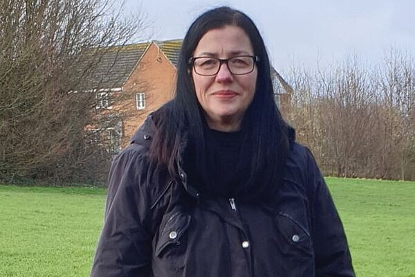 Head and shoulders picture of Morag Everden with glasses and dark shoulder length hair in a park with buildings and trees in the background.