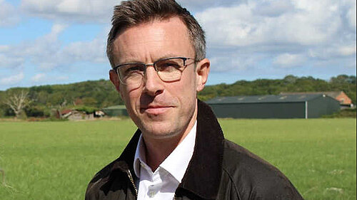 A close up of Ben Dempsey, an open field behind him and farm buildings in the background