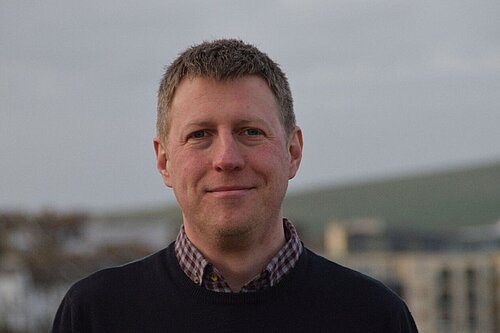 James MacCleary head and shoulders against blurred background of buildings and sky